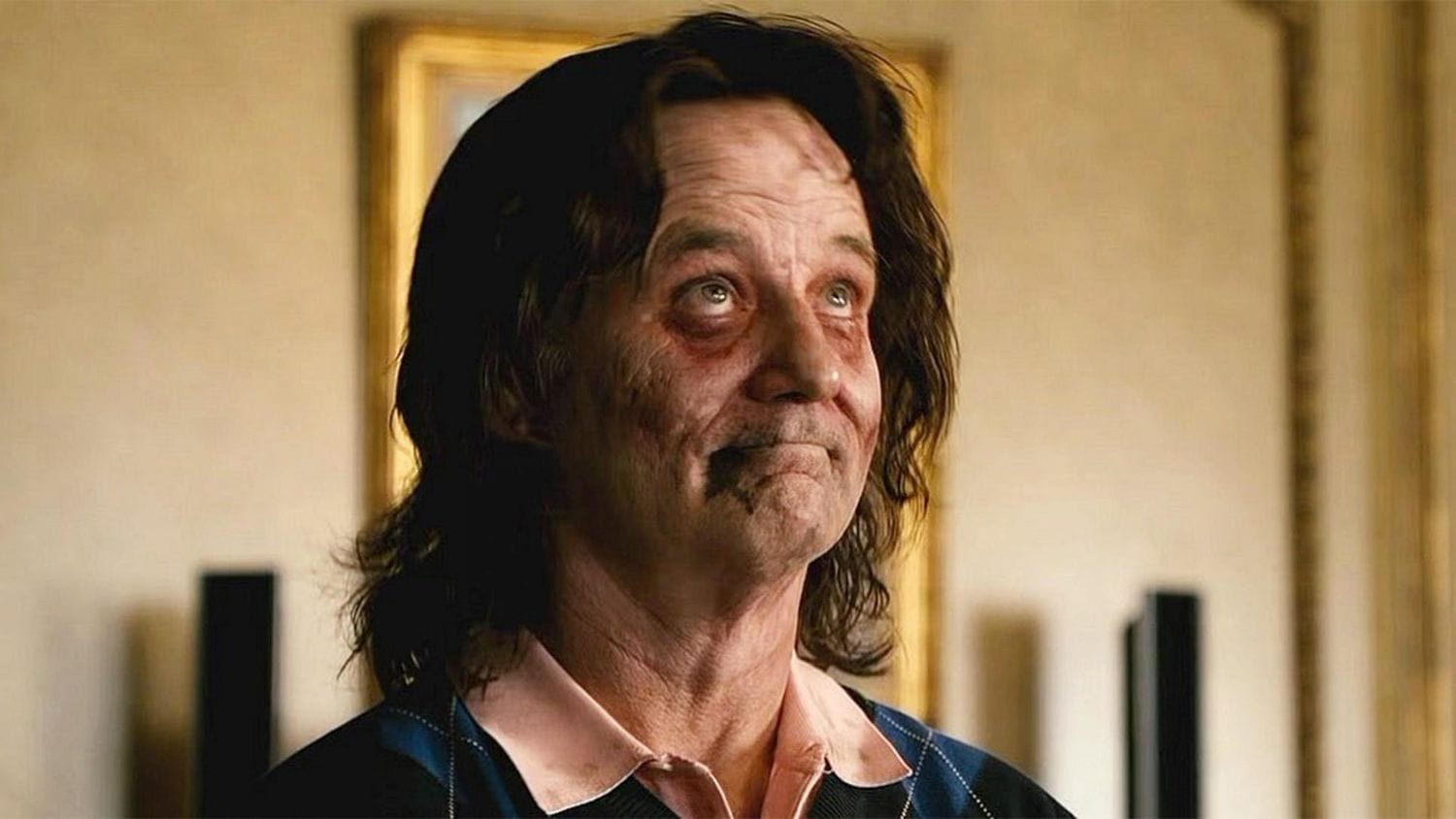 A still from "Zombieland" (2009) showing actor Bill Murray in a close-up shot. He appears disheveled with long, messy dark hair and makeup that makes him look undead or zombie-like, with dark circles around his eyes. He's wearing a blue jacket with a light-colored shirt and has a subtle, characteristically wry expression on his face. The scene is lit with warm, golden tones, and there's a blurred beige wall with framed artwork in the background.